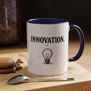 Può includere: Una tazza in ceramica bianca con interno e manico blu scuro. La parte anteriore presenta la parola INNOVATION in lettere maiuscole nere sopra un disegno al tratto in bianco e nero di una lampadina accesa con raggi e una piccola stella.