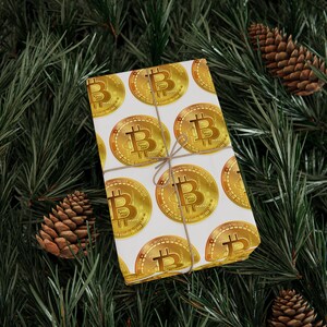 May include: A rectangular gift box wrapped in white paper featuring a repeating pattern of gold Bitcoin symbols. The box is tied with thin, light brown twine and is positioned against a background of dark green pine needles and brown pinecones.