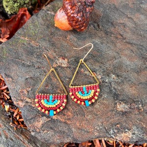 May include: A pair of dangle earrings featuring a triangular gold-tone metal frame with a semi-circular beaded section. The beads are arranged in concentric rows of red, yellow, and turquoise, creating a vibrant pattern suspended from a hook.
