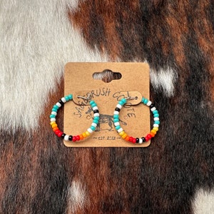 May include: A pair of turquoise, white, orange, red, and black beaded hoop earrings. The earrings are on a brown card with the text "Sagebrush Coyote est. 2022" printed on it.