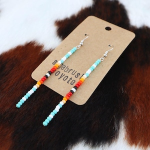 May include: A pair of long beaded earrings with turquoise, white, orange, red, and black beads. The earrings are hanging from a brown card with the text "Sagebrush Coyote" on it.