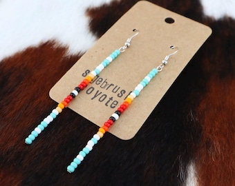 May include: A pair of long beaded earrings with turquoise, white, orange, red, and black beads. The earrings are hanging from a brown card with the text "Sagebrush Coyote" on it.