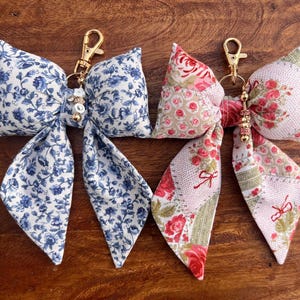 May include: Two fabric bow keychains with gold metal rings and beaded charms. One bow is blue and white floral print, the other is pink and white floral print.