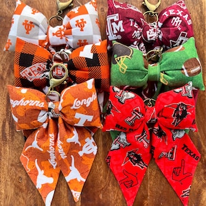 May include: A collection of fabric bow keychains in various colors and patterns. The bows feature collegiate team logos and mascots, including the Texas Longhorns, Texas A&M, and others. Each bow has a gold-colored metal clasp.