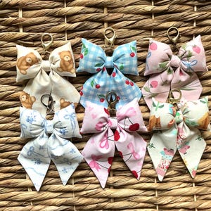 May include: A collection of fabric bow keychains with gold-colored hardware. The bows feature various patterns, including teddy bears, cherries, strawberries, and floral designs. The keychains are arranged on a woven wicker background.