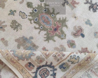 May include: Beige area rug with a detailed pattern in shades of blue, green, and red. The rug has a folded corner, revealing a border with a geometric design. The overall design is intricate and traditional, suitable for home decor.