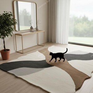 Puede incluir: Una alfombra moderna con un diseño abstracto en tonos grises, marrones y crema. Un gato negro camina sobre la alfombra. La alfombra está colocada sobre un suelo de madera en una habitación con un espejo y una consola.