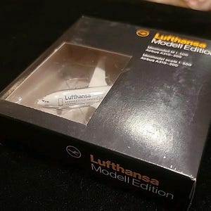 May include: A Lufthansa Modell Edition miniature airplane model in a black box. The model is a white Airbus A310-300, visible through a clear plastic window. The box has the Lufthansa logo and model details printed on it.