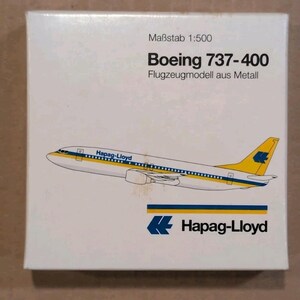 May include: A white box featuring a Boeing 737-400 model airplane with yellow and blue accents. The text on the box reads "Maßstab 1:500" and "Flugzeugmodell aus Metall". The Hapag-Lloyd logo is also visible.