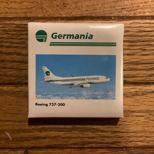 Può includere: Un piccolo quadrato bianco con il logo Germania e una striscia verde. L'immagine presenta un aereo Boeing 737-300 che vola in un cielo blu. Il logo Germania è un cerchio verde con un aereo bianco.