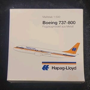 May include: A white box featuring a Boeing 737-800 model airplane. The model is white with orange and blue accents, and the Hapag-Lloyd logo is visible. Text on the box includes "Maßstab 1:500" and "Flugzeugmodell aus Metall."