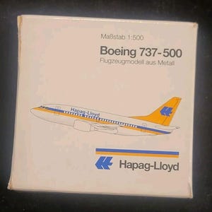 May include: A white box featuring a Boeing 737-500 model airplane with the Hapag-Lloyd logo. The airplane is white with yellow and blue accents. The box also includes the text "Maßstab 1:500" and "Flugzeugmodell aus Metall."