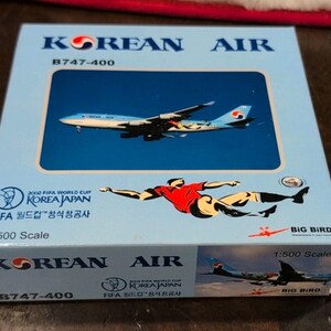 以下が含まれることがあります： 韓国航空B747-400の飛行機モデルが入った水色の箱。箱には「2002 FIFAワールドカップ韓国日本」の文字とサッカー選手のグラフィックが入っています。モデルは1:500スケールです。