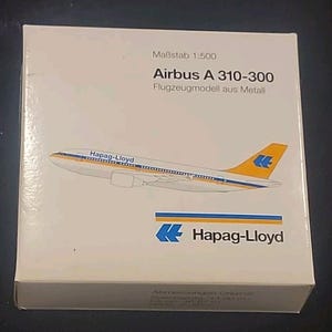 May include: A box featuring a model airplane. The box has the text "Airbus A 310-300" and "Flugzeugmodell aus Metall". The airplane is white with yellow and blue accents and the "Hapag-Lloyd" logo.