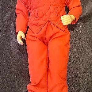 May include: A vintage action figure of a humanoid alien in a red quilted uniform and black boots. The figure has a green reptilian head with detailed facial features. The outfit includes a quilted vest and matching pants.