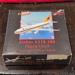May include: A boxed model airplane of an Airbus A310-300, branded as Hapag Lloyd. The model is white with orange and blue accents. The box is black with text and a picture of the plane. The scale is 1:500.