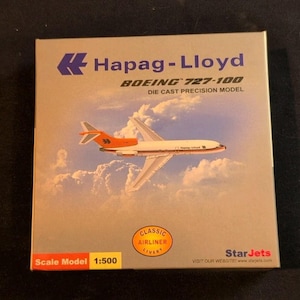 May include: A die-cast precision model of a Boeing 727-100 airliner in a box. The box features the Hapag-Lloyd logo and the words "BOEING 727-100". The model is a scale model 1:500, with a classic airliner livery.