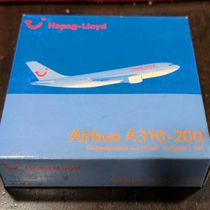 May include: A blue box featuring the Hapag-Lloyd logo and an illustration of an Airbus A310-200 airplane. The text "Airbus A310-200" is printed in orange. The box is a model airplane.