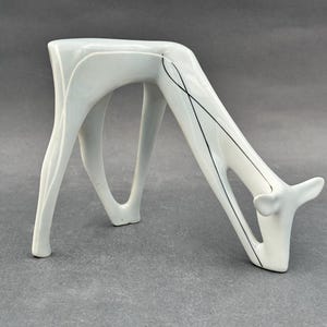 May include: A white ceramic deer sculpture in a drinking posture. The sculpture has a modern design with black accent lines. The deer's legs form the base, and the head is lowered as if drinking. A decorative item.