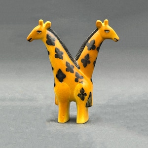 Może przedstawiać: Ceramiczna figurka przedstawiająca dwie głowy żyrafy przymocowane do jednego korpusu. Rzeźba jest jaskrawożółta z ciemnobrązowymi, czteropłatkowymi plamami w kształcie kwiatów na szyjach i tułowiu oraz ciemną grzywą wzdłuż każdej szyi.
