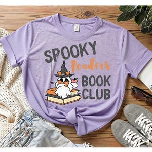 May include: A light purple t-shirt featuring a graphic of a ghost wearing a witch hat and reading a book. The text reads "SPOOKY Readers BOOK CLUB" in black and orange lettering. The ghost is sitting on a stack of books with a small spider hanging nearby.