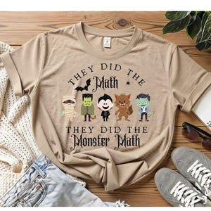 May include: A tan short-sleeved t-shirt featuring a graphic of five cartoon monsters and the text "THEY DID THE MATH" and "THEY DID THE MONSTER MATH". The monsters include a mummy, Frankenstein's monster, a vampire, a werewolf, and a zombie.
