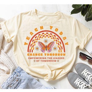 May include: A cream-colored short-sleeve t-shirt featuring a graphic print of a butterfly, daisies, and a checkered rainbow. The text reads: TEACH TODAY CHANGE TOMORROW EMPOWERING THE LEADERS OF TOMORROW in orange and red lettering.