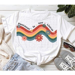 May include: A white short-sleeve t-shirt featuring a graphic print of wavy horizontal stripes in red, orange, yellow, and teal. The text "PROGRESS is NOT LINEAR" is printed in black above the stripes, accented by small colorful flower illustrations.