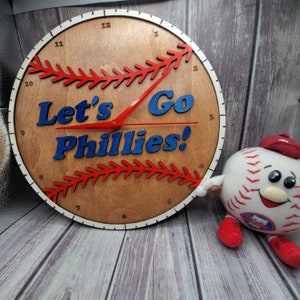 May include: A wooden baseball-themed clock with the words "Let's Go Phillies!" in blue. The clock has red stitching details and hands, with a white border. A baseball mascot plush toy is to the right.