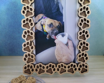May include: A wooden picture frame with a paw print design. The frame is holding a photo of a brown and white dog. The dog is wearing a harness and is looking at the camera. There are dog biscuits on a plate in the foreground.