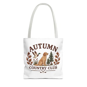 Può includere: Una borsa tote bianca con una stampa grafica che riporta il testo AUTUMN COUNTRY CLUB. Il design include un cane dorato seduto, un germano reale in volo, due pini, foglie di quercia e due mazze da golf incrociate con una pallina da golf.