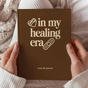 Puede incluir: Un diario de tapa dura color marrón oscuro con el texto en color crema "in my healing era" acompañado de un icono de corazón y un gráfico de tirita, con el texto "trust the process" impreso en una fuente más pequeña en la parte inferior.