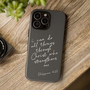 May include: A dark gray smartphone case featuring the white handwritten text "I can do all things through Christ who strengthens me" followed by "Philippians 4:13". The case has a cutout for a triple-lens camera system on the upper left side.