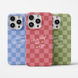 May include: Three phone cases with a checkered pattern in blue, red, and green. Each case has a different handwritten quote: "slow down you're doin fine", "life is tough but so are you", and "what's the best that could happen?"