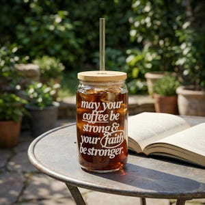 May include: A clear glass coffee tumbler filled with iced coffee, a bamboo lid, and a glass straw. The tumbler has white text that reads "May your coffee be strong & your faith be stronger."