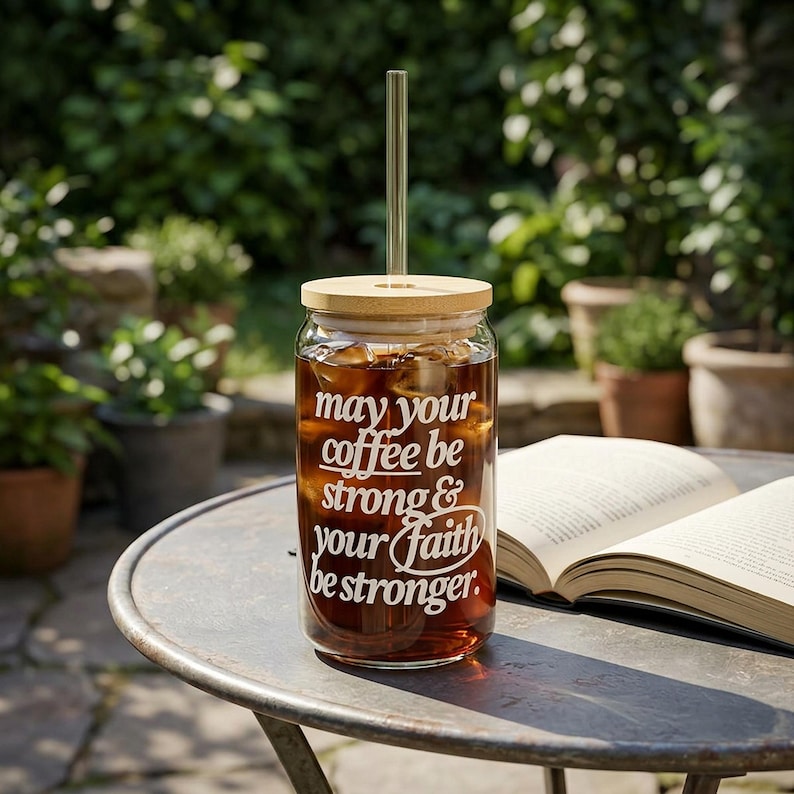 May include: A clear glass coffee tumbler filled with iced coffee, a bamboo lid, and a glass straw. The tumbler has white text that reads "May your coffee be strong & your faith be stronger."