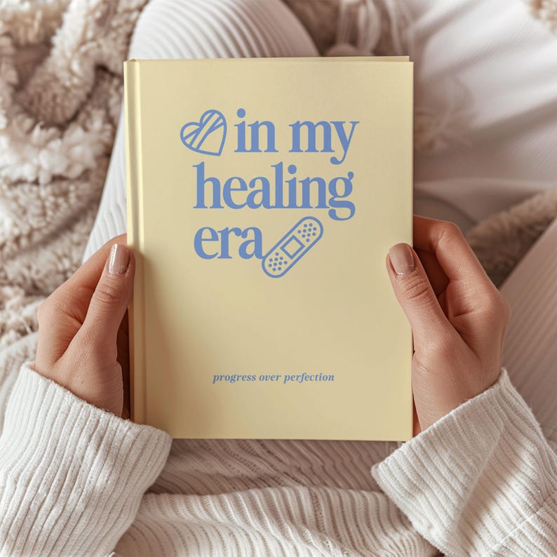 May include: A pale yellow journal with the words "in my healing era" in blue script, along with a heart and bandage graphic. The phrase "progress over perfection" is printed at the bottom.