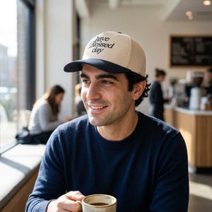 A man with dark hair wears a cream and black baseball cap embroidered with "have a Blessed day" and holds a coffee cup in a cafe.