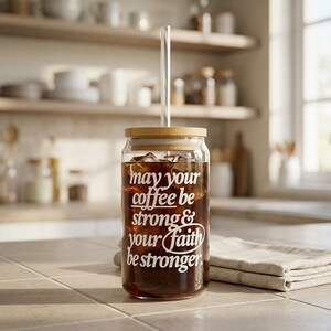 May include: A clear glass coffee tumbler with a wooden lid and a glass straw. The tumbler is filled with iced coffee and features the white text, "may your coffee be strong & your faith be stronger."