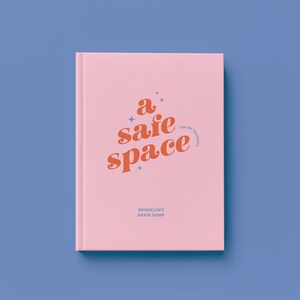 May include: A pink journal with the words "a safe space" in orange script. The text "for my thoughts" is in a smaller font. Below, the text "Brooklyn's Brain Dump" is printed in a smaller font. The journal is on a blue background.