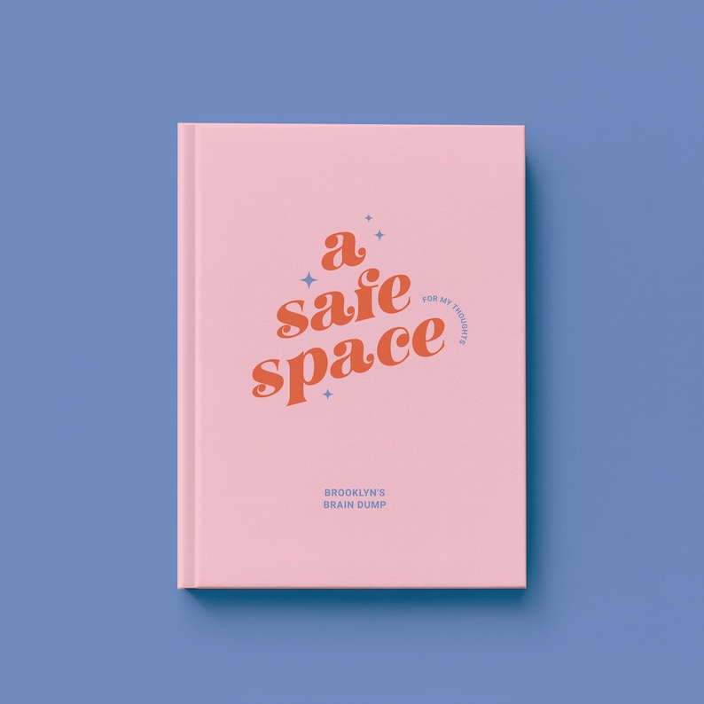 May include: A pink journal with the words "a safe space" in orange script. The text "for my thoughts" is in a smaller font. Below, the text "Brooklyn's Brain Dump" is printed in a smaller font. The journal is on a blue background.