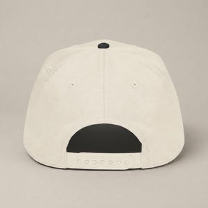 The back of a cream-colored baseball cap with a black button on top and an adjustable white plastic snap closure.