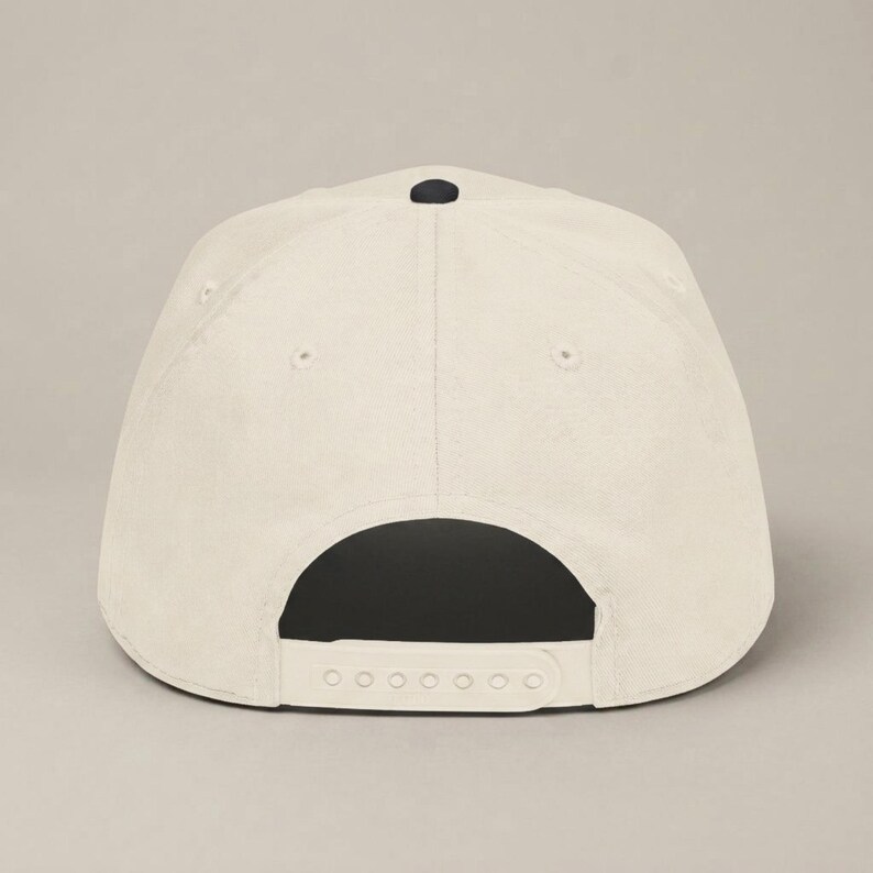 The back of a cream-colored baseball cap with a black button on top and an adjustable white plastic snap closure.
