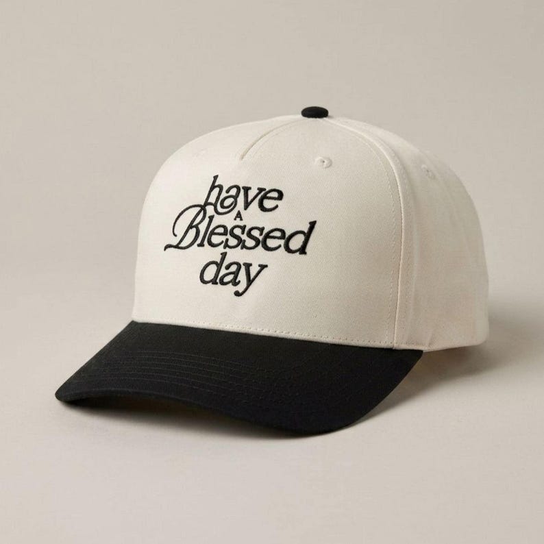 A two-tone baseball cap with a black brim and a cream crown, embroidered with the words "have a Blessed day" in black script, viewed from a slightly different angle.