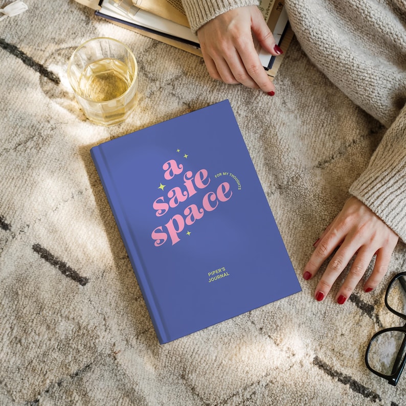 May include: A blue journal with the words "a safe space" in pink script. The journal also has the text "for my thoughts" and "Piper's Journal". The journal is on a patterned rug.