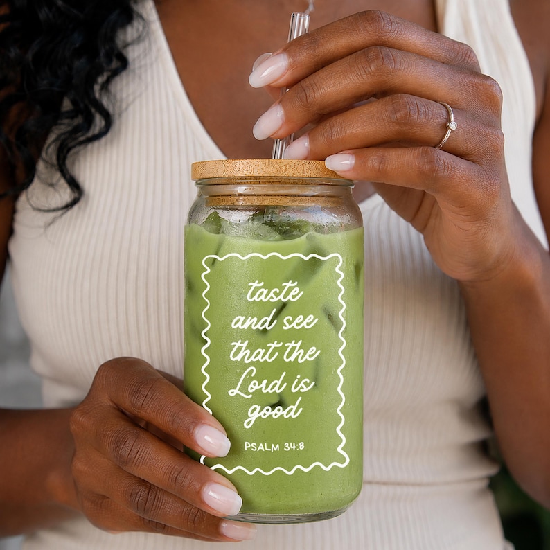May include: A clear glass jar with a wooden lid, filled with a green beverage and ice cubes. The jar has a white rectangular label with the text "taste and see that the Lord is good" and "PSALM 34:8". A clear straw is inserted into the jar.