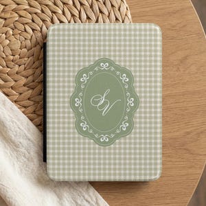 May include: A rectangular e-reader case featuring a light sage green and white gingham pattern. Centered on the front is a sage green oval emblem with a white decorative scrollwork border, containing the white cursive monogram letters SV.