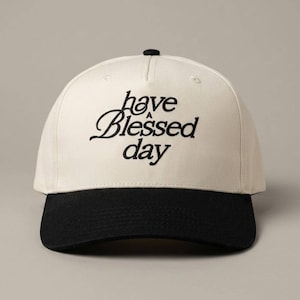 A two-tone baseball cap with a black brim and a cream crown, embroidered with the words "have a Blessed day" in black script.