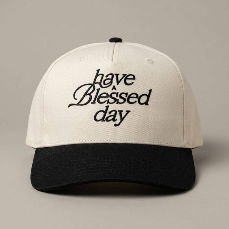 A two-tone baseball cap with a black brim and a cream crown, embroidered with the words "have a Blessed day" in black script.