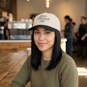A woman with dark hair wears a cream and black baseball cap embroidered with "have a Blessed day" while sitting at a wooden table in a cafe.